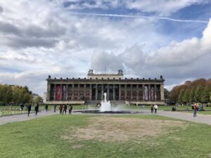 A large, classical building is fronted by many columns and topped with two statues of horses, a large fountain spouts water in the middle of a patchy looking lawn, visitors stroll along the concrete paths. The sky above has heavy cloud with patches of blue, trees line the sides of the lawns.