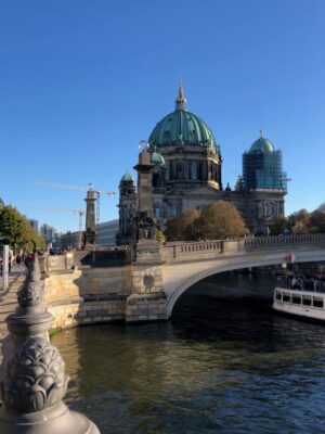 The blue green dome of a cathedral is seen across a wide river bridge under a bright blue sky while flat riverboats pass beneath the bridge.