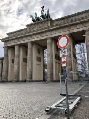 A red and white street sign and metal fencing stands in front of the grand sandstone columns of the Brandenburg Gate, the four horses pull a chariot atop the grand archway.