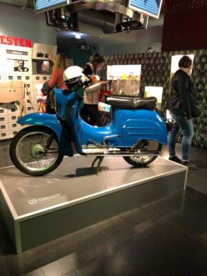 A blue, compact scooter stands pride of place in a museum, complete with a white helmet resting on top, brightly lit up, visitors browse the exhibits behind it