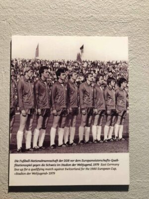 A small black and white picture is displayed of members of the DDR soccer team in their team kit lined up before a game, adhered to a grey plaster wall.