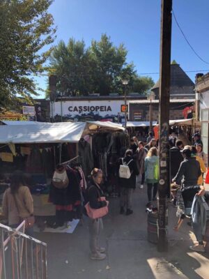 Shoppers browse flea market stalls set up under a blue sky, a sign on an adjacent building reads Cassiopeia and trees dot the square.