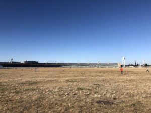 A large, golden field stretches back into the distance, ringed by a semi-circular terminal building, once an airport, now a sporting field for Berlin. A small white tower is seen to the right of the old terminal building under the blue sky.