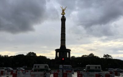 The Victory column in Berlin, topped with a golden statue, stands tall against a stormy sky as the sun is setting, vans and barricades close off the road.