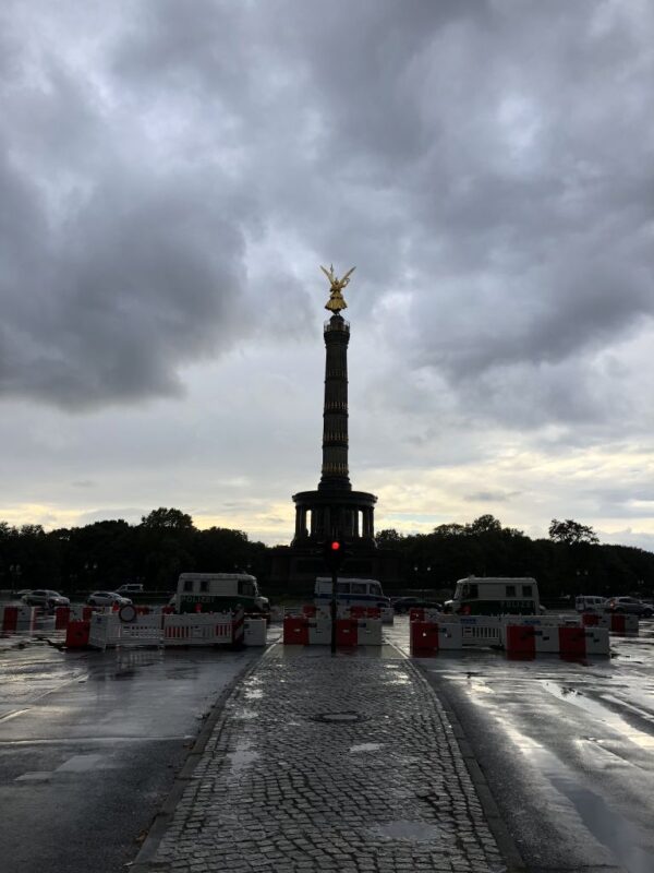 The Victory column in Berlin, topped with a golden statue, stands tall against a stormy sky as the sun is setting, vans and barricades close off the road.