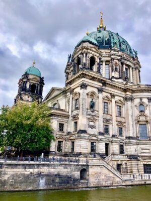 The light stonework and blue-green copper domes of the Berlin Cathedral are contrasted against a stormy sky and green Spree river flowing past.