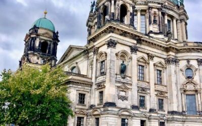 The light stonework and blue-green copper domes of the Berlin Cathedral are contrasted against a stormy sky and green Spree river flowing past.