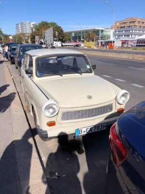 A vintage cream car with stark white rings around its headlights, known as a Trabant is parked parallel to the footpath on a city street in Berlin, the sky above is blue.