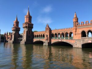 A red brick and grey stone bridge stretches across a brown river, complete with turrets and battlements and large arcade windows which offer views to the blue sky behind, somewhat reminiscent of a castle's features.