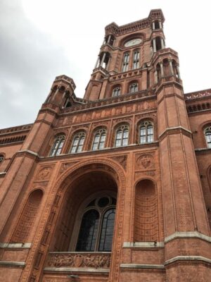 A bold, red bricked tower stretches to a heavily clouded sky, ornate windows and dramatic columns frame the tower as it ascends, complete with a large clock face on its anterior.