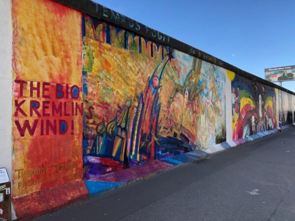 Rainbow coloured murals decorate the concrete expanse of the former Berlin Wall, including one saying the big Kremlin wind, a billboard stands over the wall against a blue sky. Enjoy more sights like this in this Berlin 3 day itinerary.