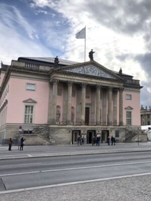 A large, powder pink building with multiple stone columns completing its facade is topped by classical statues and gold lettering. The grand city street is wide and lined with neoclassical buildings, the sky is building with thunderclouds.