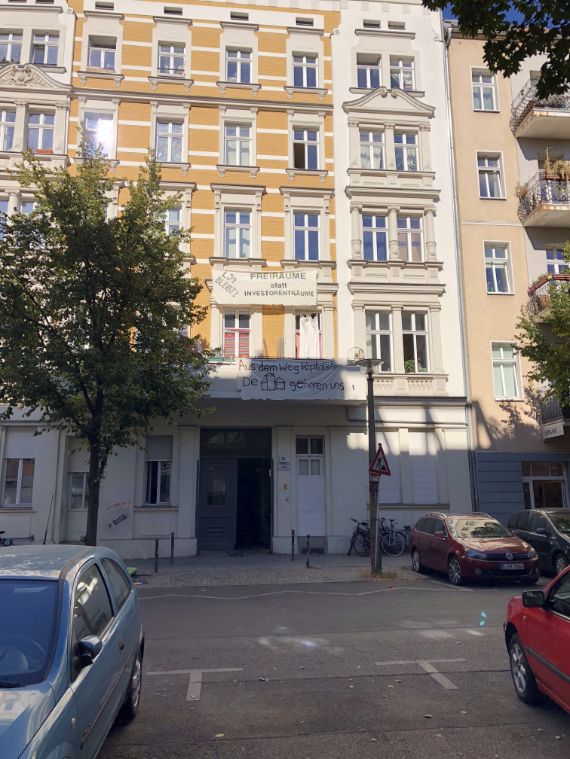 A city apartment block is shown, painted a light golden-brown with white accents, reaching over 5 stories high is plastered with political signs, cars are parked before the row of apartment buildings which is partially in sun.