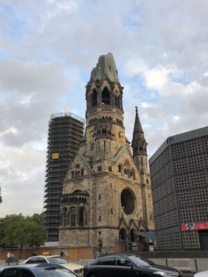 A partially ruined sandstone church with the lower portion of the tower remaining stands preserved with futuristic city buildings behind it, traces of green copper still line the decorative elements of its rooflines and there is a large hole where a stained glass rose window would have once rested.