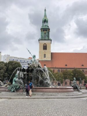 A red-roofed cathedral, complete with cream tower and copper green spire stands behind an ornate fountain featuring Neptune on a cobblestone square. Visitors mill around the base of the fountain under a clouded sky