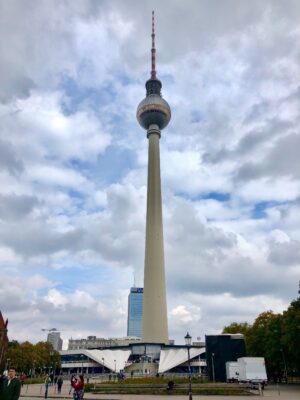 A large, concrete, beige tower topped with a silver sphere and red and white antenna stretches up to the clouded sky atop a large city square.