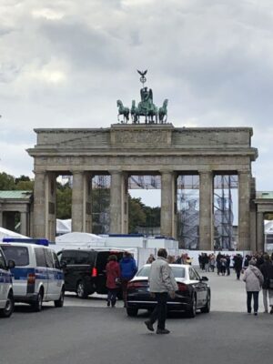 The sandstone, neo-classical gateway of the Brandenburg Gate stands at the end of Unter den Linden, multiple vans and gathered people crowd the foreground, the four copper horses atop the gate are clearly visible.