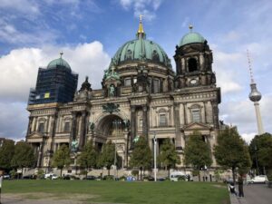 A large, copper domed cathedral stands on a grassy square, the blue green domes are topped with golden orbs and crucifixes. A spherical restaurant and red and white spike top the Television Tower in the background, the sky behind them is blue and dusted with clouds.