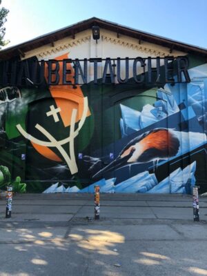 A large, colourful mural of a bird and abstract geometric designs is painted onto the wall of an old warehouse, the sky above is blue and the ground is paved with concrete