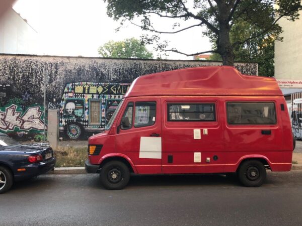 A red, vintage van is parked on the side of a dark street, the mural painted on the adjacent wall also features a van.