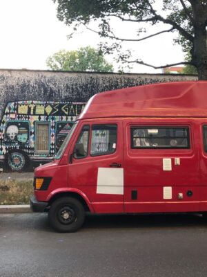 A red, vintage van is parked on the side of a dark street, the mural painted on the adjacent wall also features a van.