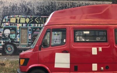 A red, vintage van is parked on the side of a dark street, the mural painted on the adjacent wall also features a van.