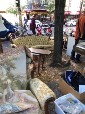 Yellow and white vintage furniture is set up in a park in the centre of Berlin, including glassware in a plastic tub and shoppers browsing the market.