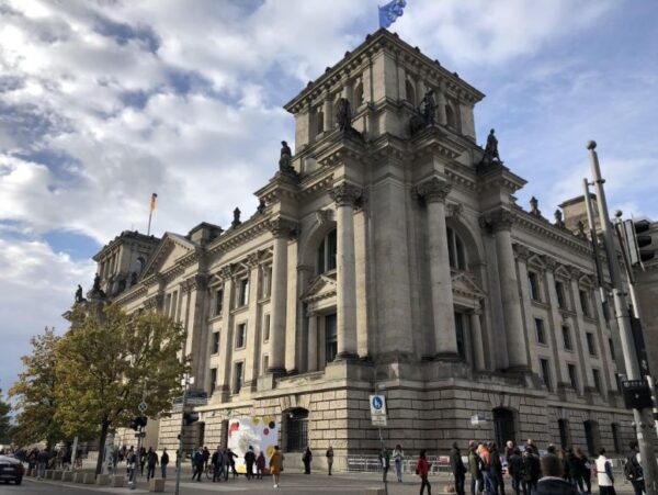 A large, imposing stone building with statues on each corner and German and EU flags flying above, people mill on the footpaths surrounding the buildings and a blue sky is dusted with clouds above.