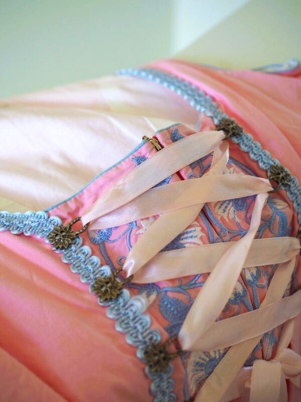 The bodice of a pink, traditional dirndl dress is seen, with metallic eyelets designed to resemble flowers attached to decorative blue trim and a pink ribbon running between them.