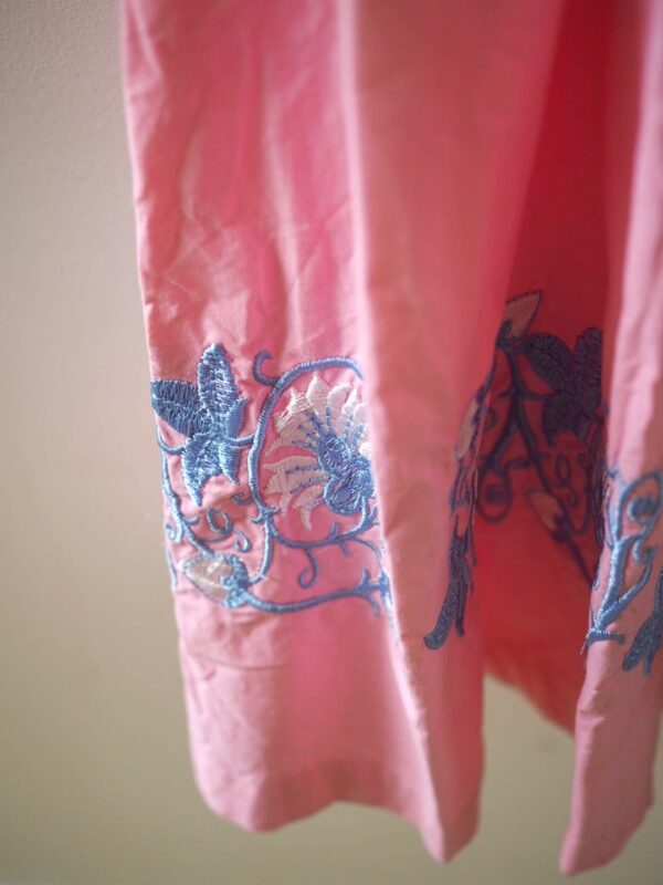 Crinkled pink fabric forms the skirt of a dirndl, embroidered with floral and vine motifs in blue and white along the hemline against a cream wall.