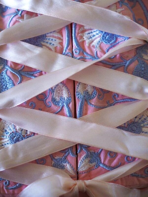 A pink ribbon is seen criss-crossing a pink bodice with a zip running down the middle and intricate blue and white embroidery in floral patterns.