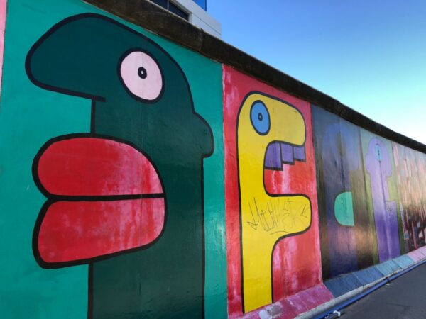 Large murals featuring humanoid caricatures in different colours are painted onto a remaining section of the Berlin Wall, blue sky is seen above the wall.