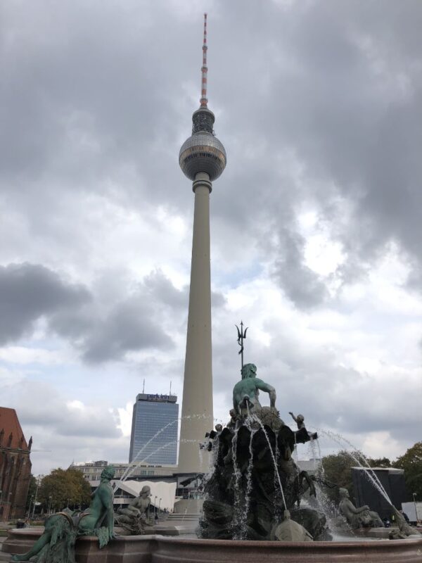 A large, spike topped tower rises into the dark clouds with a shiny spherical section in the middle. Beneath, a fountain gushes with coppery green figures, including a man with a trident.