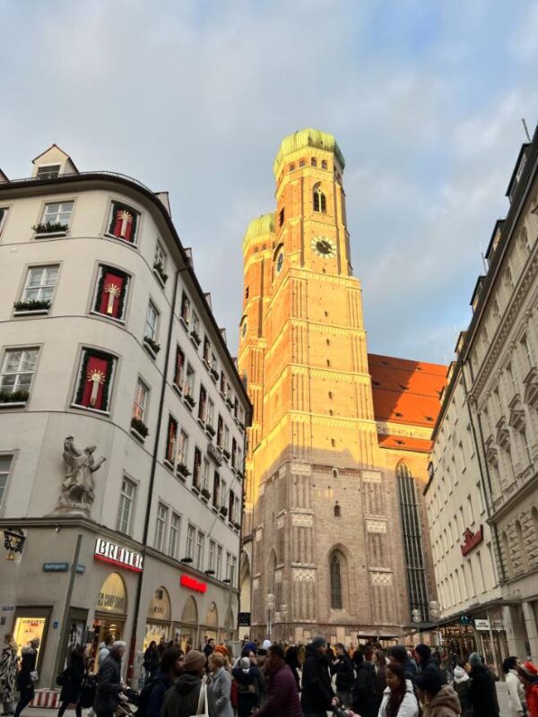 Two large, brick towers stand tall against the sky in the middle of Munich's Old Town while visitors hurry here and there below, The onion shapes dome shine green in the golden light and the sky above has mostly clouded over.