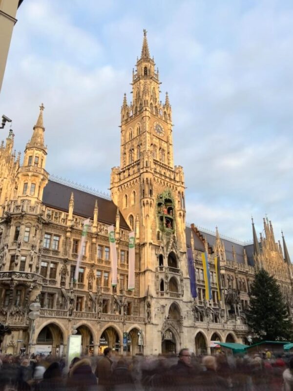 Crowds mill about under the fairytale spires of the neo Gothic Neues Rathaus in Munich, the last hints of daylight have turned the rooftop golden and the sky above has some cloud. A Christmas tree also stands in the square.