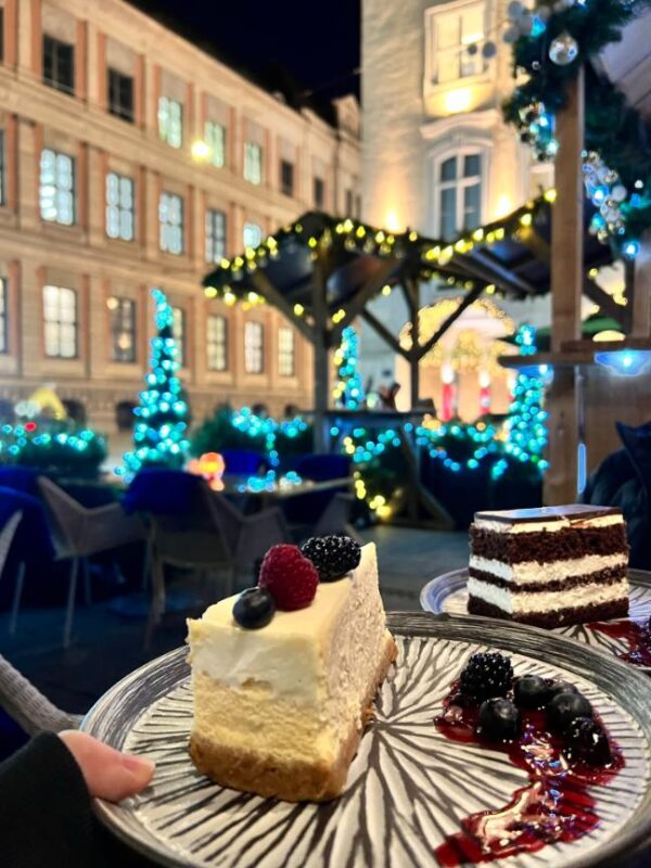 A hand holds up a plate of cheesecake topped with berries, accompanied by berries and sauce on the side of the striped, grey plate, a second plate of chocolate cake with cream is seen slightly behind it while Christmas lights show in the background.