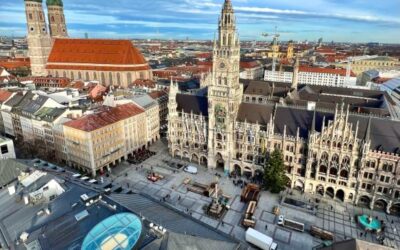 A rooftop view over the old town of Munich is seen with the many spired New Town Hall and the red roofed Frauenkirche with its double spires are both seen under a blue sky dusted with some cloud. The square of Marienplatz is mostly empty.