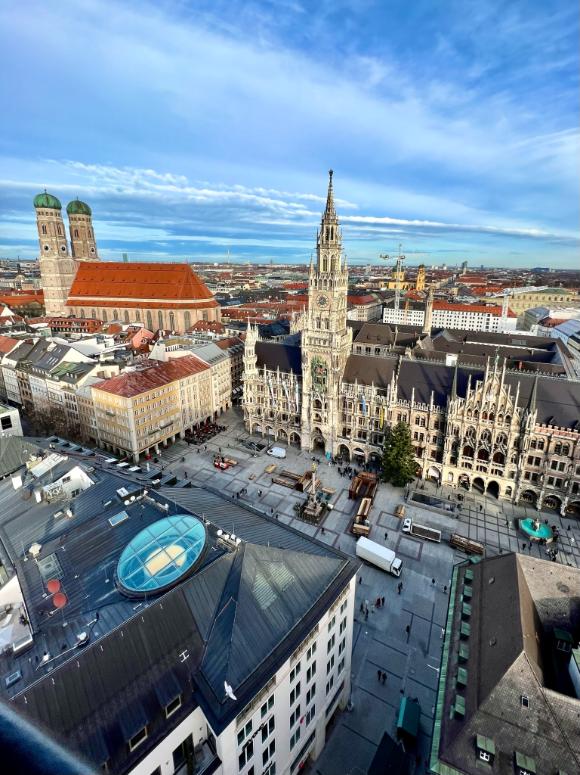 A rooftop view over the old town of Munich is seen with the many spired New Town Hall and the red roofed Frauenkirche with its double spires are both seen under a blue sky dusted with some cloud. The square of Marienplatz is mostly empty.