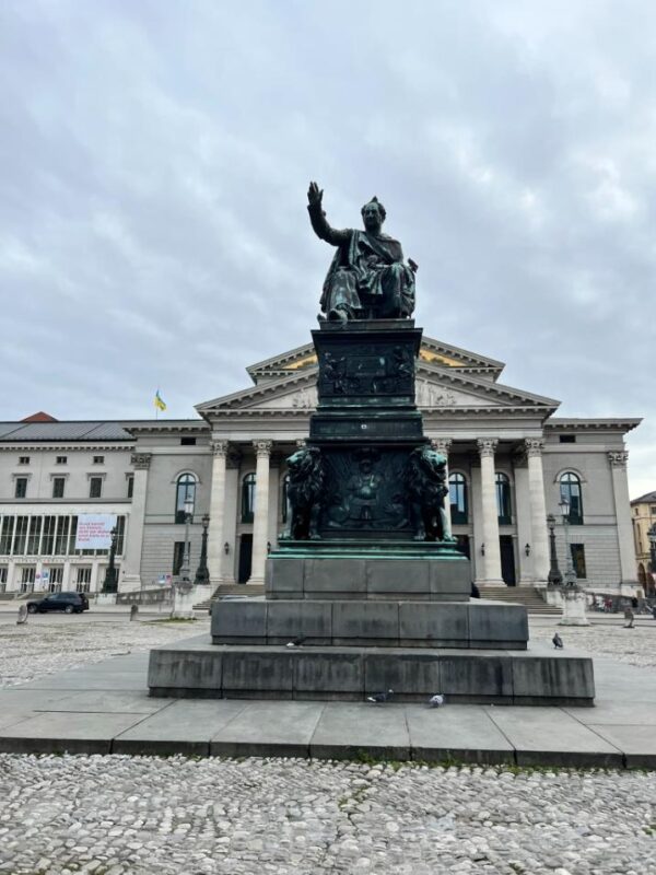 A large, dark green statue is seen standing in the centre of a city square on a large plinth. Behind it, a grand opera house is seen and the sky above is clouded over, cobblestones line the street.