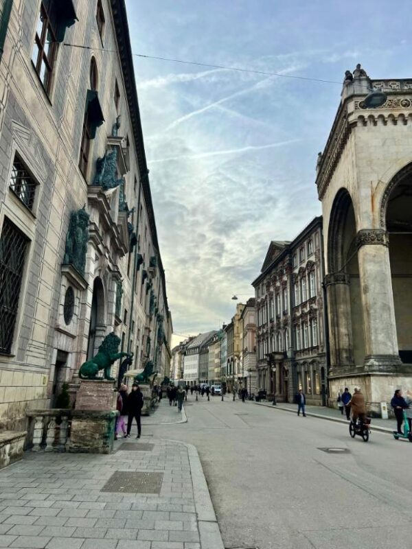 The shot is taken along a city street in Munich, lined on both sides by elegant buildings. On the left hand side, two blue-green lion statues guard a gate into the city palace, above the blue sky is crisscrossed with jet trails and light cloud.