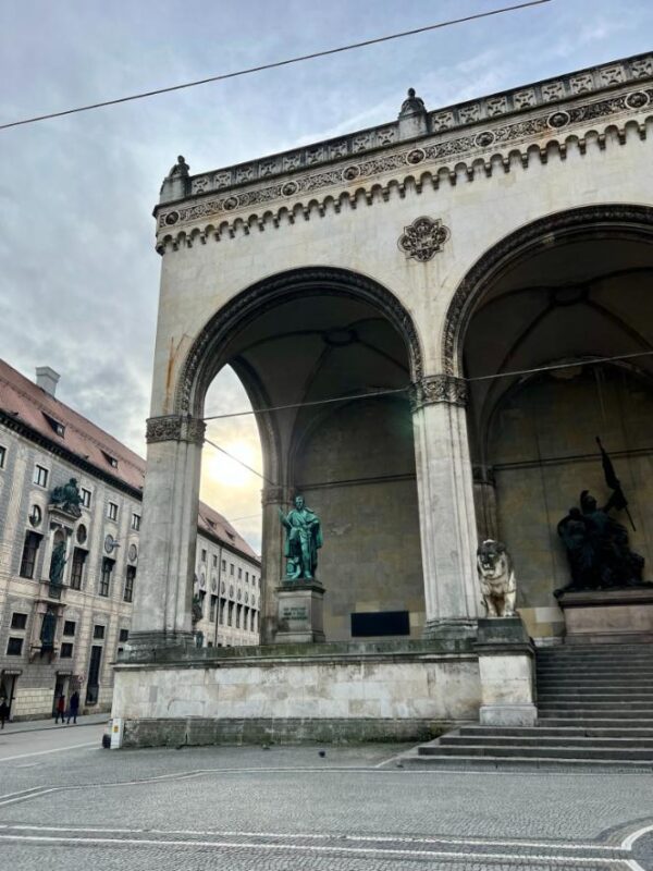 A large stone building with high archways and detailing houses statues of copper blue on a city square. The sky above is lightly clouded and grey.