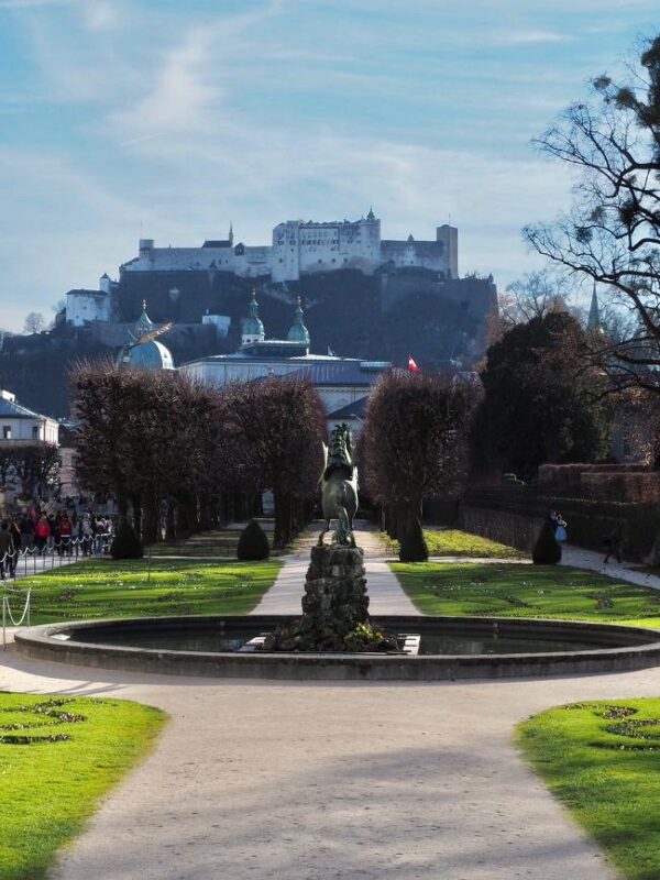 A white stone fortress rises high above manicured gardens, complete with a fountain topped with a horse statue. Multiple copper domes stand in the distance.