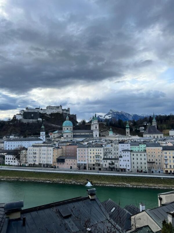 A city scene is shown from across a wide, green river, pastel townhouses line the banks while multiple copper tipped spires are seen rising above the roofs of the Old Town. Above the scene, a large white fortress stands high on the hill, beneath a darkly clouded sky