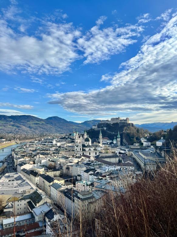 A large city is seen from an elevated perspective, with multiple buildings made out of white stone, including churches with copper green domes stand in the valley under a blue sky dusted with cloud. At the far end of the valley, a large white fortress rises high on a rocky outcrop and hills reach into the background.