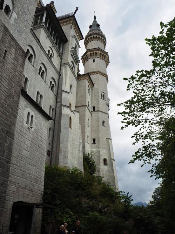 Light coloured stone walls and turreted towers stretch up towards a cloudy sky, greenery surrounds the righthand edge of the image. Small rounded windows dot the walls and towers. Enjoy these views on a day trip from Munich to Neuschwanstein Castle