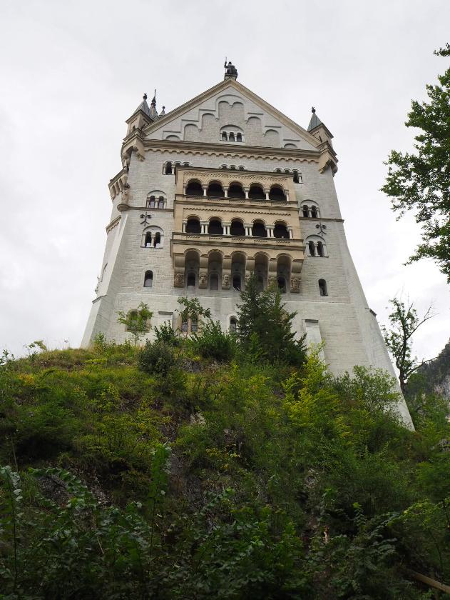 The large end of a castle, complete with multiple balconies painted gold stretches up to the cloudy sky, perched on a greenery covered slope. Small, decorative towers cover each corner while there is decorative while trim on the very top story and a statue on the roof.