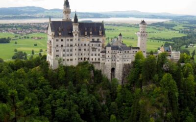 A large, white castle topped with multiple dark roofed turrets and towers stands against a backdrop of green fields and lakes, surrounded by greenery