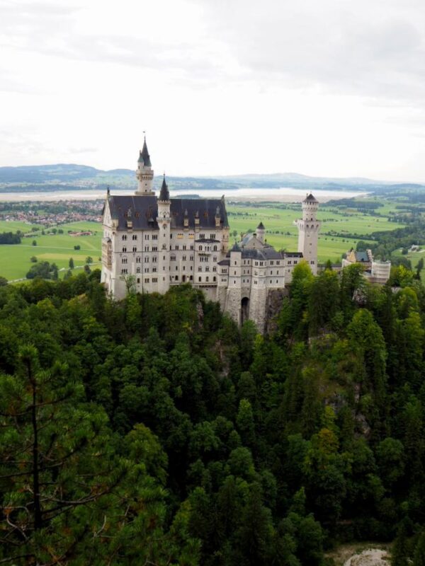 A large, white castle topped with multiple dark roofed turrets and towers stands against a backdrop of green fields and lakes, surrounded by greenery