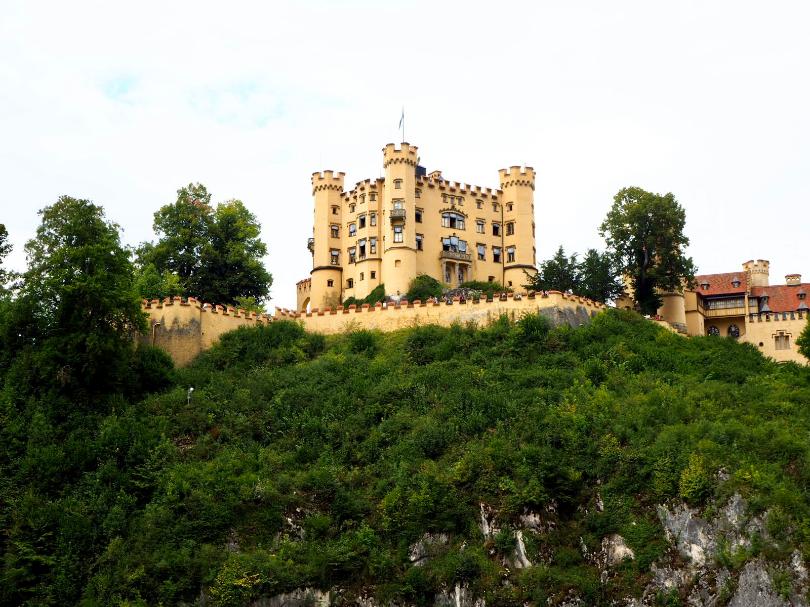 A pale yellow castle stands against a cloudy sky surrounded by green, leafy trees and a matching yellow wall, topped with terracotta tiles. Towers stand at each of the castles' corners, shrubs runs down the slope towards the viewer.