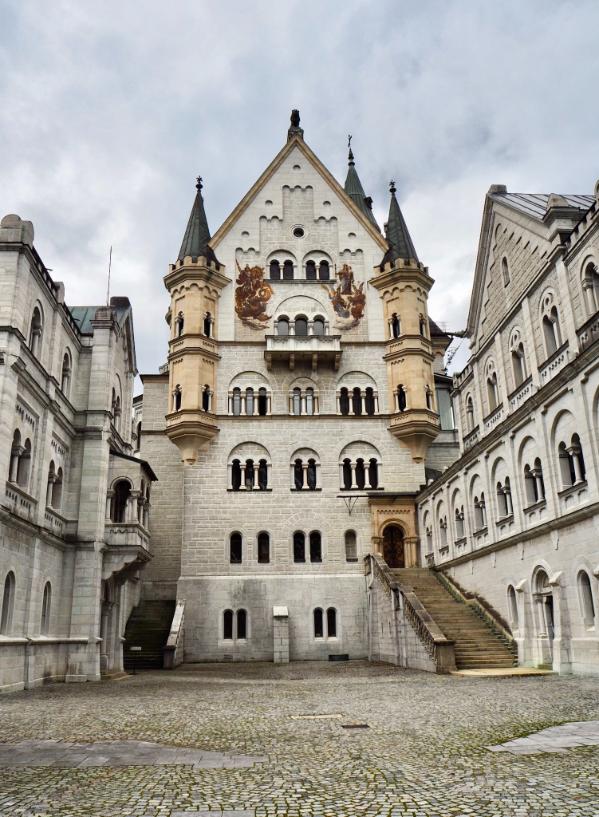 A large, cobblestoned courtyard is surrounded on all sides by large castle walls peppered with windows. Centrally, a large edifice faces the viewer with double golden turrets and heraldic animals, as well as decorative edges around the top stories, the sky above is thickly clouded